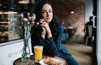 Delighted woman in stylish wear sitting at wooden round table and enjoying fresh juice and cookie during breakfast in cafe